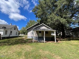 A small, single-story house with a covered front porch and steps, situated on a grassy lawn with large trees nearby. Other homes are visible on either side under a partly cloudy sky.