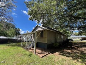 A single-story beige house with a covered front porch, steps leading to the entrance, and a large tree providing shade in the yard. The sky is partly cloudy and neighboring houses are visible in the background.