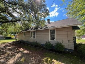 Single-story house with light-colored siding, several windows, and a dark shingled roof. The house is surrounded by trees and shrubs, with sunlight filtering through the branches.