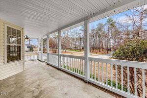 A spacious covered front porch with a white railing, concrete floor, and a ceiling light, overlooking a quiet suburban street with trees and dry grass visible beyond the yard.