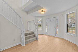 A bright entryway with light wood flooring, white walls, a white front door with glass panels, three large windows, and a staircase with white railings and carpeted steps.