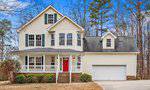 Charming two-story beige house at 94 Oak Forest Drive, Henderson, with green shutters, a red front door, a front porch, an attached two-car garage, and a dormer window above the garage. Surrounded by trees and a patchy lawn.