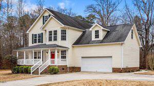 Two-story beige house with black roof, red front door, covered porch, and attached two-car garage. The driveway is wide, and the yard has dry grass and a few shrubs. Trees without leaves are in the background.