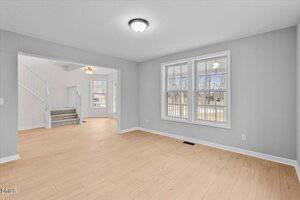 A bright, empty room with light wood flooring, gray walls, a ceiling light fixture, and two large windows facing outside. An open doorway leads to a staircase and another well-lit room.