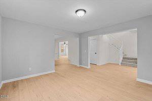 A bright, empty room with light wood floors, light gray walls, a ceiling light fixture, visible doorways leading to a staircase, and adjoining spaces. No furniture or decorations are present.