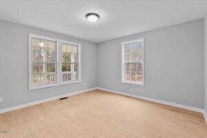 Empty room with light wood flooring, gray walls, white trim, and a white ceiling with a central light fixture. Two large windows let in natural light and show an outdoor view with trees and a porch railing.
