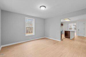 A bright, empty room with light wood flooring, light gray walls, a window on the left, and an overhead light fixture. The room opens into a kitchen area with an island and a door leading outside.
