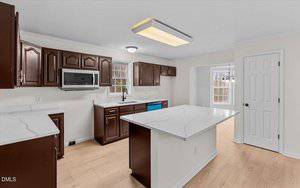 A modern kitchen with dark brown cabinets, white marble countertops, light wood floors, a center island, built-in microwave, and large windows letting in natural light. A dining area is visible through an open doorway.