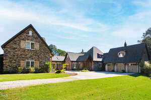A large stone house with multiple peaked roofs, a three-car garage, and shuttered windows sits on a spacious green lawn under a clear blue sky. A gravel path runs alongside a concrete driveway.