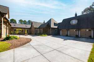 Large stone and wood house with multiple gables, three-car garage, and spacious concrete driveway on a sunny day. Some landscaping and greenery are visible along the edges of the driveway.