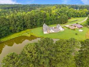 Aerial view of a large house with multiple gables and an attached garage, situated next to a pond and surrounded by dense trees and greenery, with a driveway leading to a detached structure.