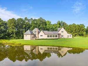 A large, cream-colored house with multiple gables and a gray roof sits on a grassy lawn by a pond, with trees in the background and the house reflected in the still water.