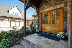 A stone walkway leads to double wooden front doors with glass panes, surrounded by brick and stone walls. White potted flowers sit on either side of the entrance. A detached garage is visible in the background.