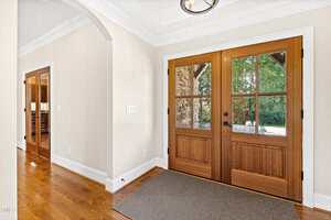 A spacious entryway with wooden double doors featuring glass panels, a rectangular doormat on hardwood floors, arched doorways, and neutral-colored walls. Trees and greenery are visible through the doors.