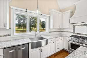 A modern white kitchen features a farmhouse sink, stainless steel appliances, marble countertops, and a large window overlooking a green landscape with trees. White cabinets and a tile backsplash complete the space.