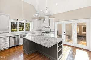 Spacious modern kitchen with marble countertops, a large central island, stainless steel appliances, white cabinets, wood flooring, and double glass doors leading to a patio. Sunlight streams in through the windows.