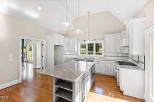 A spacious, bright kitchen with white cabinets, stainless steel appliances, a large gray island with marble countertop, pendant lights, and hardwood floors. There are windows above the sink and a door to the right.