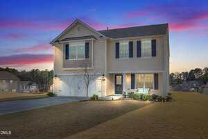 Two-story beige house with dark shutters at 2802 W Brookwood Court, Creedmoor, featuring an attached double garage and small front porch. Exterior lights glow as the home sits on a large grassy lot at dusk under a colorful sky.