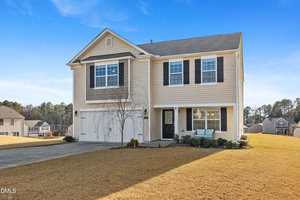 Two-story beige house with black shutters, attached two-car garage, and a small front porch with seating. The house is surrounded by a dry, yellowed lawn and has a clear blue sky in the background.