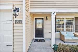 Front porch view of a beige house with black door, wall-mounted lanterns, house number 2802, window with seating area, and a green garden hose on the ground.
