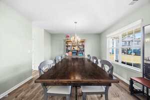 A dining room with a long wooden table surrounded by eight gray chairs. A chandelier hangs above the table, and a bookshelf filled with board games stands against the far wall. Sunlight streams through a large window.