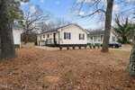 A single-story beige house at 215 Wilmington Avenue in Oxford, featuring black shutters and a small front porch, is surrounded by trees and a yard covered in pine needles. Another green-sided house sits in the background beneath a clear sky.