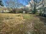 A grassy, partially cleared lot at 219 Alexander Avenue, Oxford features tall, dry grass in the foreground, scattered trees, a small white shed, and several objects including a large tank and tarp-covered items under a blue sky.