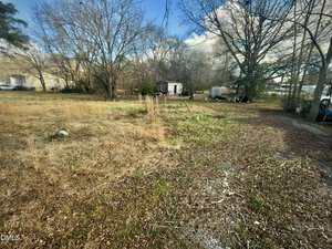 A grassy, partially cleared lot at 219 Alexander Avenue, Oxford features tall, dry grass in the foreground, scattered trees, a small white shed, and several objects including a large tank and tarp-covered items under a blue sky.