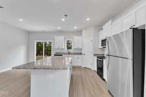 Modern kitchen with white cabinets, stainless steel appliances, granite countertops, a large island, light wood flooring, and a pantry door. Sliding glass doors in the background open to a view of trees.