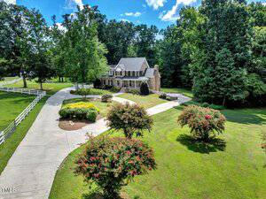 The two-story brick house at 1086 Roberts Chapel Road, Stem, features white trim and is nestled at the end of a curved driveway, surrounded by green lawns, landscaped shrubs, and mature trees under a blue sky with scattered clouds.
