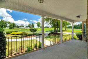 View from a covered porch with white columns and black railing, looking out onto a landscaped yard with shrubs, a walkway, green grass, trees, and white fencing under a partly cloudy sky.