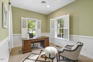 A home office with green walls, two windows with white shutters, a wooden desk with a chair, a modern armchair, a white ottoman, and a beige rug with brown patterns on a carpeted floor.