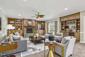 A spacious living room with built-in wooden shelves, a stone fireplace, and several gray and beige sofas and chairs arranged around a large dark coffee table. Windows and glass doors let in natural light.