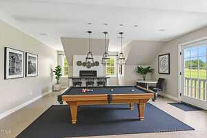 A modern game room with a pool table on a black rug, three pendant lights above, a seating area with two chairs and a round table, wall art, a fireplace, and a door leading outside. Bright and neutral decor.