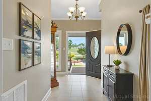 A view of a home entryway with beige walls, a tile floor, four abstract paintings on the left, a console table with a lamp and plant on the right, a round mirror above, and an open door showing a lawn outside.