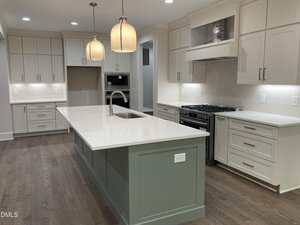 A modern kitchen with light wood floors, white cabinetry, a large island with a sink, stainless steel appliances, two pendant lights above the island, and neutral tile backsplash.