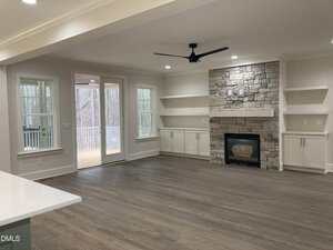 A spacious living room with light-colored walls and hardwood floors, featuring a stone fireplace with built-in shelves and cabinets on both sides, large windows, and sliding doors leading to a covered porch.
