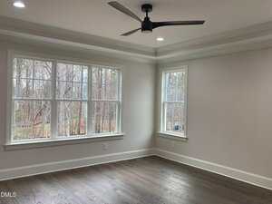 An empty room with light-colored walls, large windows showing trees outside, a dark ceiling fan, and wood flooring. Recessed lighting is on in the ceiling.