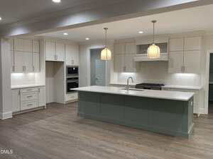 A modern kitchen with white cabinets, a large green island with a white countertop, double wall ovens, a built-in stove, pendant lights, and wood flooring. The space is well-lit with recessed and pendant lighting.