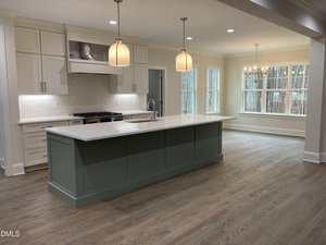 Modern kitchen with white cabinets, a green island with a sink, pendant lights, light wood floors, and an adjacent dining area with large windows and a chandelier.