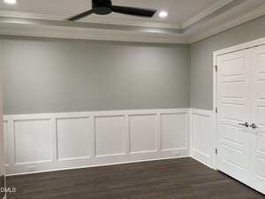 A room with light gray walls and white wainscoting, dark wood flooring, a white double door with silver handles, and a black ceiling fan with recessed lighting.
