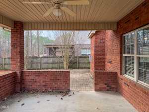 Covered brick patio with a ceiling fan, bordered by a partial brick wall. The patio overlooks a fenced backyard with bare trees and a neighboring brick house in the background. Leaves are scattered on the ground.