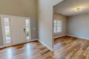 The image shows a home interior with an entryway and an adjacent room. The space has light beige walls, wood-style flooring, a large window, and a modern ceiling light fixture. A white front door is visible.