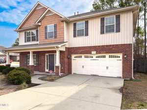A two-story house with a brick and siding exterior, covered front porch, black front door, and a two-car white garage. The driveway is concrete, and there are bushes in the front yard.