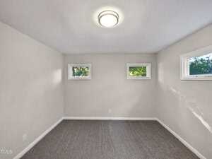 A small, empty room with light-colored walls, gray carpet, a ceiling light fixture, and three rectangular windows letting in natural light.