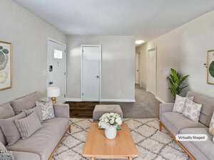 A living room with two light gray sofas, a wooden coffee table with a white flower arrangement, a white table lamp, decorative pillows, a green plant, and a white entry door visible in the background.