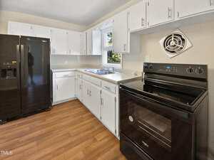 A kitchen with white cabinets, a black refrigerator, a black electric stove, a double sink under a window, and wood-look flooring. A small wall vent is above the stove. The walls are light-colored.
