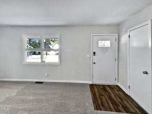 A room with light-colored walls and carpet, featuring a white door with a window, a closet door, and a double window. The entry area has wood-style flooring. Sunlight is coming through the windows.