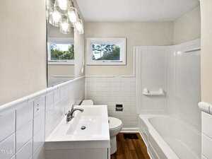 A bathroom with white tile walls, a white vanity with a sink and faucet, a toilet, a bathtub with a shower, wood-patterned floor, and two small windows above the toilet.