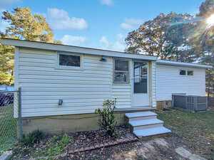 A small, white, single-story house with a flat roof, several windows, a screened door, concrete steps, and an air conditioning unit on the side, set in a partially shaded yard.